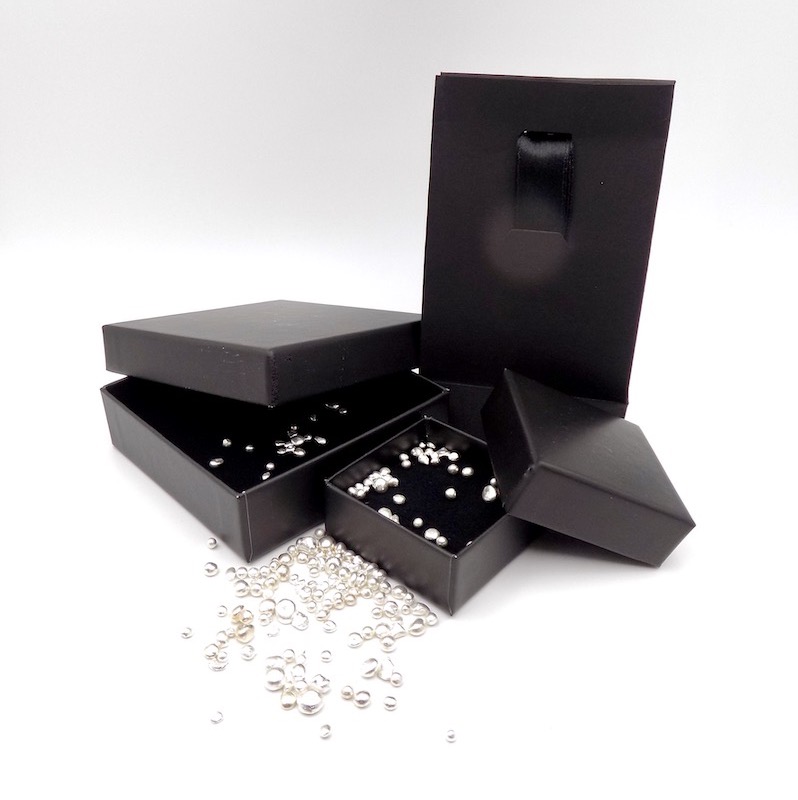 Image montrant deux écrins noirs de tailles différentes pour bijoux et un sac noir brillant. Le tout saupoudré de perles d'argent.