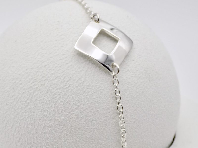 Cette image est une photo d'un bracelet en argent massif posé sur une boule blanche. Le bracelet est composé d'un carré évidé maintenu par une chaine fine en argent de part et d'autre.