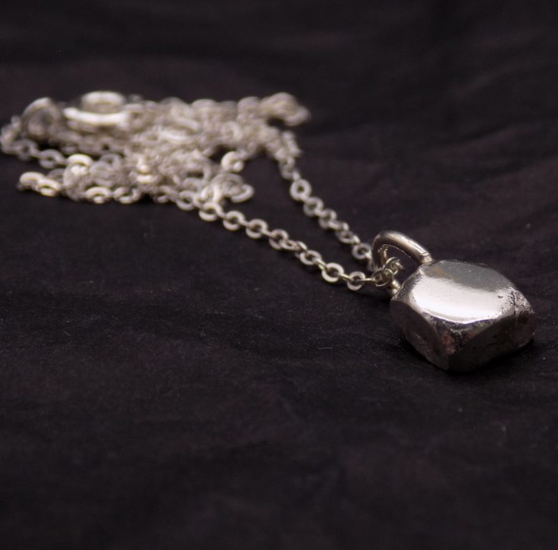 Photo d'un pendnetif en forme de pavé. il est fait tout en argent. Sur la photo il est posé sur un fond noir avec sa chaine en argent aussi.