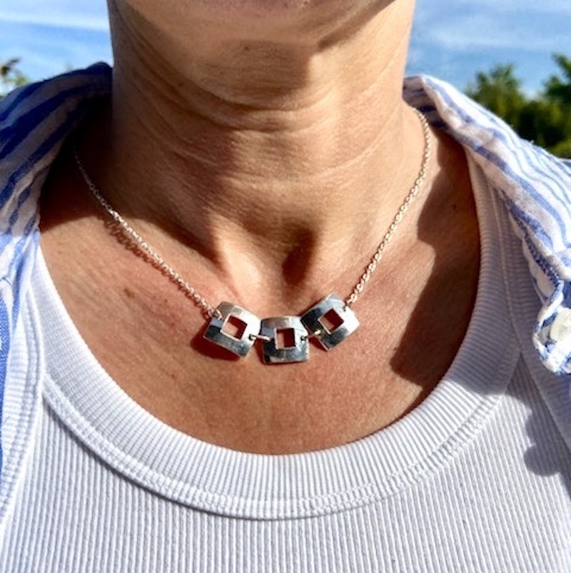 Photo en couleur d'un cou portant un collier formé de trois carrés en argent massif. La femme porte aussi un chemisier rayé balnc et bleu