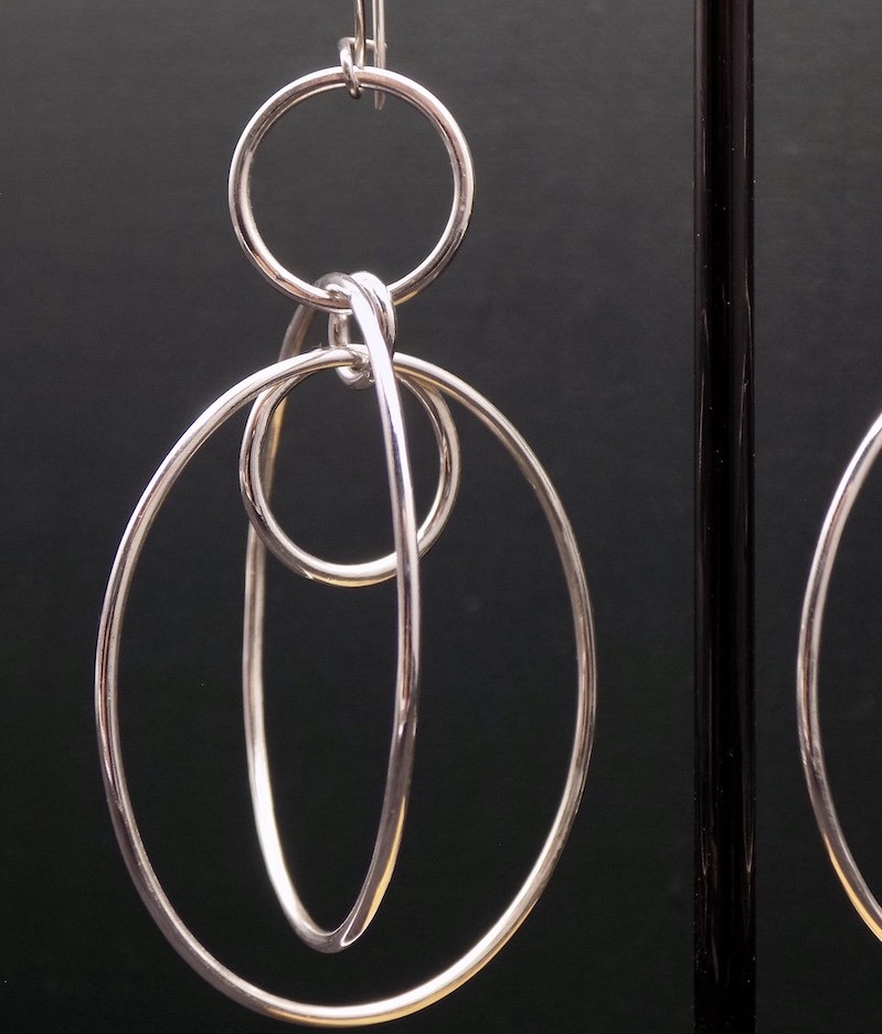 Photo d'une boucle d'oreille sur fond noir. boucle en argent réalisée à partir de fil avec plusieurs anneaux de diamètres différents qui sont entrelacés.