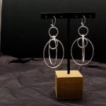 Photo un peu sombre, dans la penombre d'une paire de boucles d'oreilles longues. Les boucles sont formées de plusieurs anneaux de différentes tailles entrelacés. Boucles en argent.