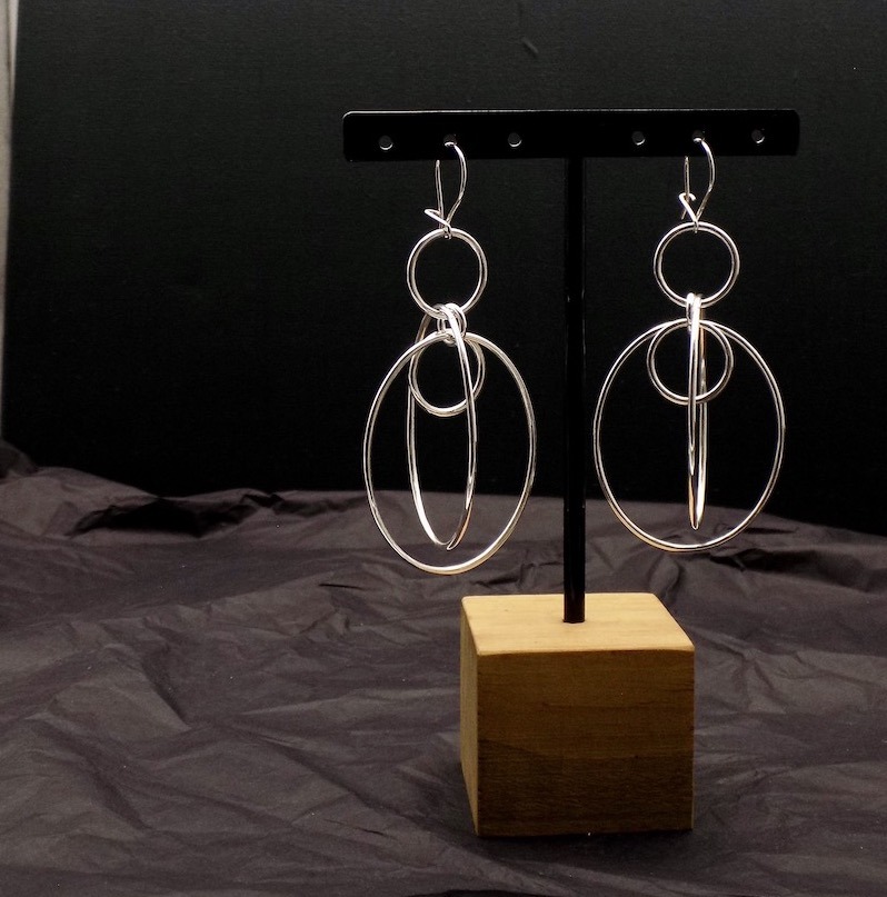 Photo un peu sombre, dans la penombre d'une paire de boucles d'oreilles longues. Les boucles sont formées de plusieurs anneaux de différentes tailles entrelacés. Boucles en argent.