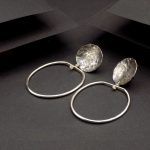 Photo vue de dessus sur un fond noir d'une paire de boucles d'oreilles tout en argent. La partie superieure est ronde martelé et oxydée puis celle du dessous est faite d'un anneau ovale.