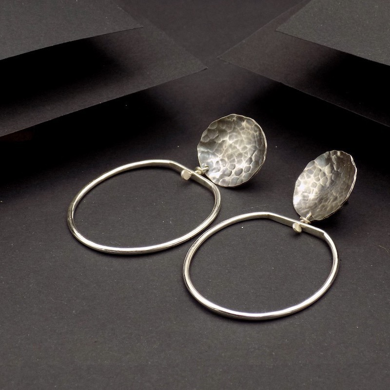 Photo vue de dessus sur un fond noir d'une paire de boucles d'oreilles tout en argent. La partie superieure est ronde martelé et oxydée puis celle du dessous est faite d'un anneau ovale.