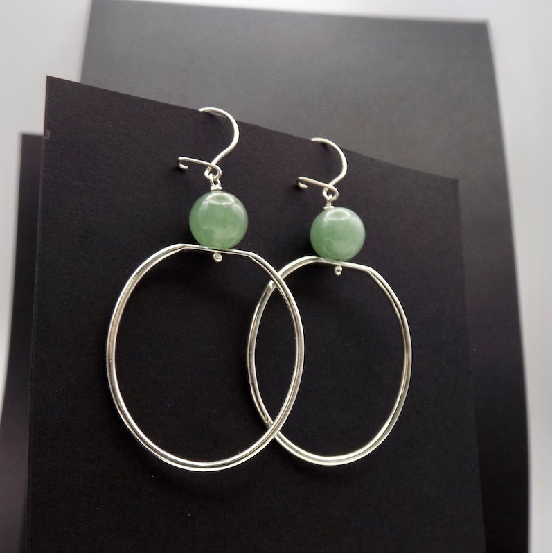 Photo d'une paire de boucles d'oreilles. Cette paire est formée de grands anneaux ovales en argent avec une perle de couleur verte claire , une pierre d'aventurine.