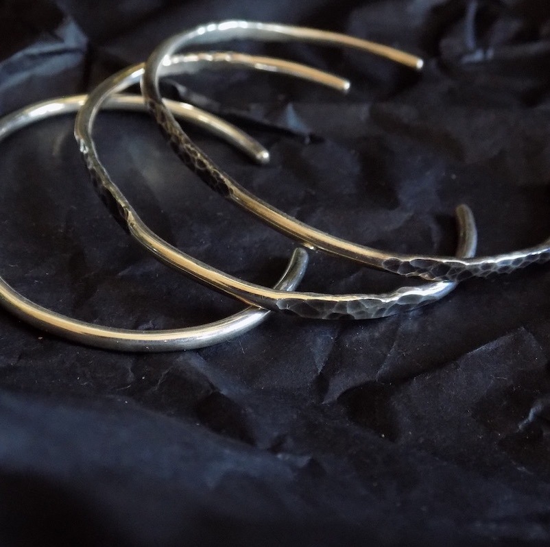 Photo sombre de trois bracelet ouverts en argent martelé. Ces bracelets sont posés les uns sur les autres et sont sur un fond noir un peu froissé.