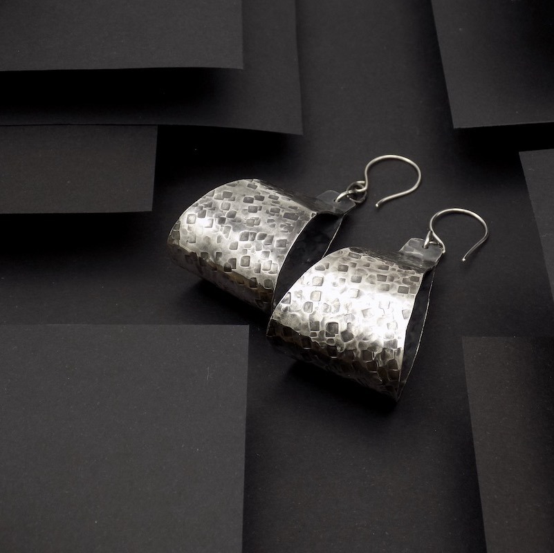 Photo sur fond noir une paire de boucles d'oreilles larges. Ces boucles sont en argent martelé puis oxydé.