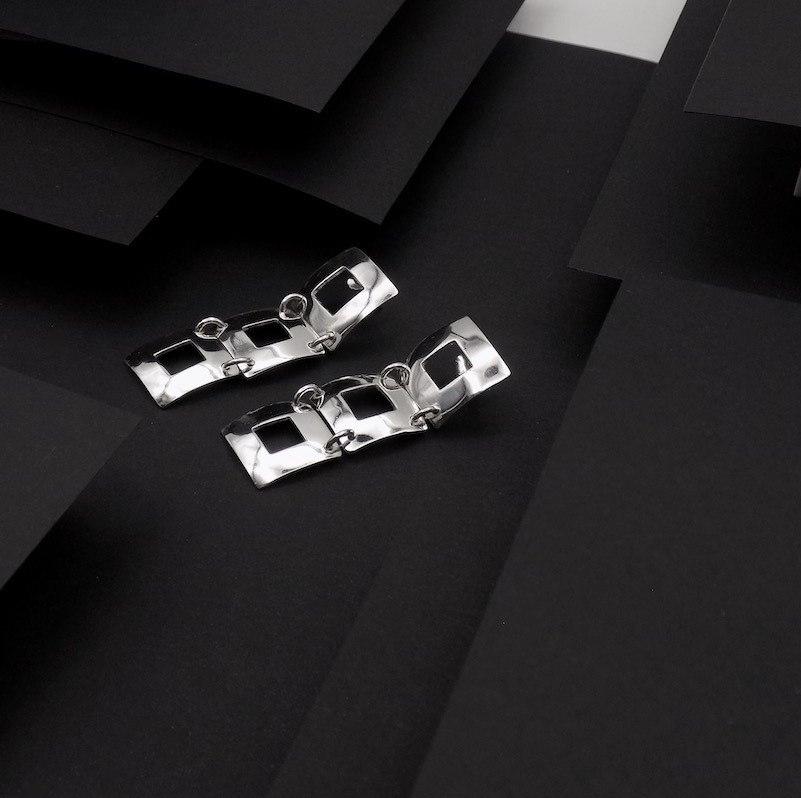 Photo faite sur fond noir pour faire ressortir une paire de boucles d'oreilles en argent. Ce sont des boucles formées de trois carrés ajourés accrochés ensemble l'un derrière l'autre